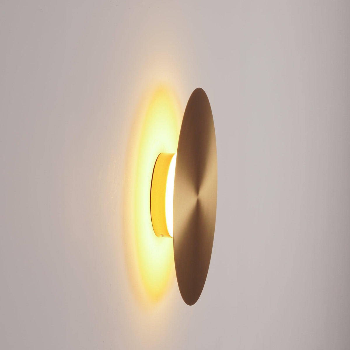 Side view of a brass Clérachic disk light lit, the amber glow shining between the disk and the pale wall behind it.