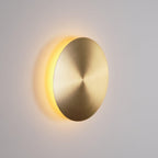 Brass Clérachic disk wall light turned slightly away, warm light glowing from behind its left edge on a grey wall.