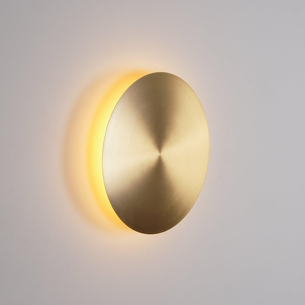 Brass Clérachic disk wall light turned slightly away, warm light glowing from behind its left edge on a grey wall.