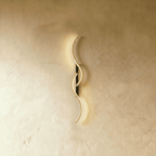 Clérachic S-shaped wall light with two interlocking gold LED curves on a slim black backplate, glowing against warm limewashed plaster.