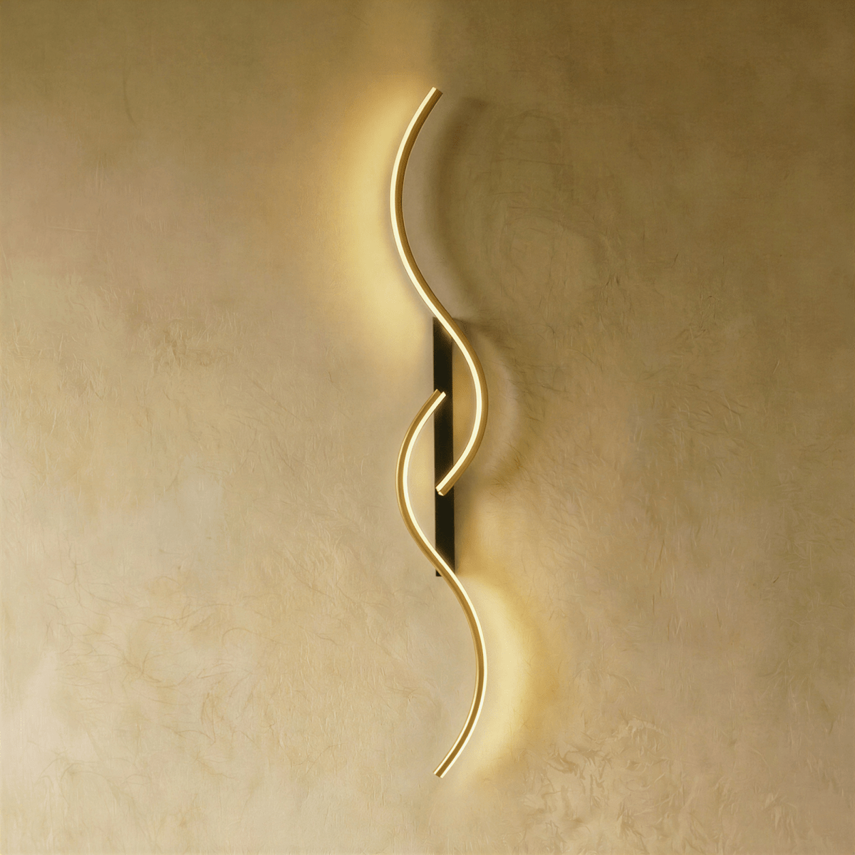 Clérachic S-shaped wall light with two linked gold-toned LED curves and a narrow black backplate, giving a warm glow on plaster.