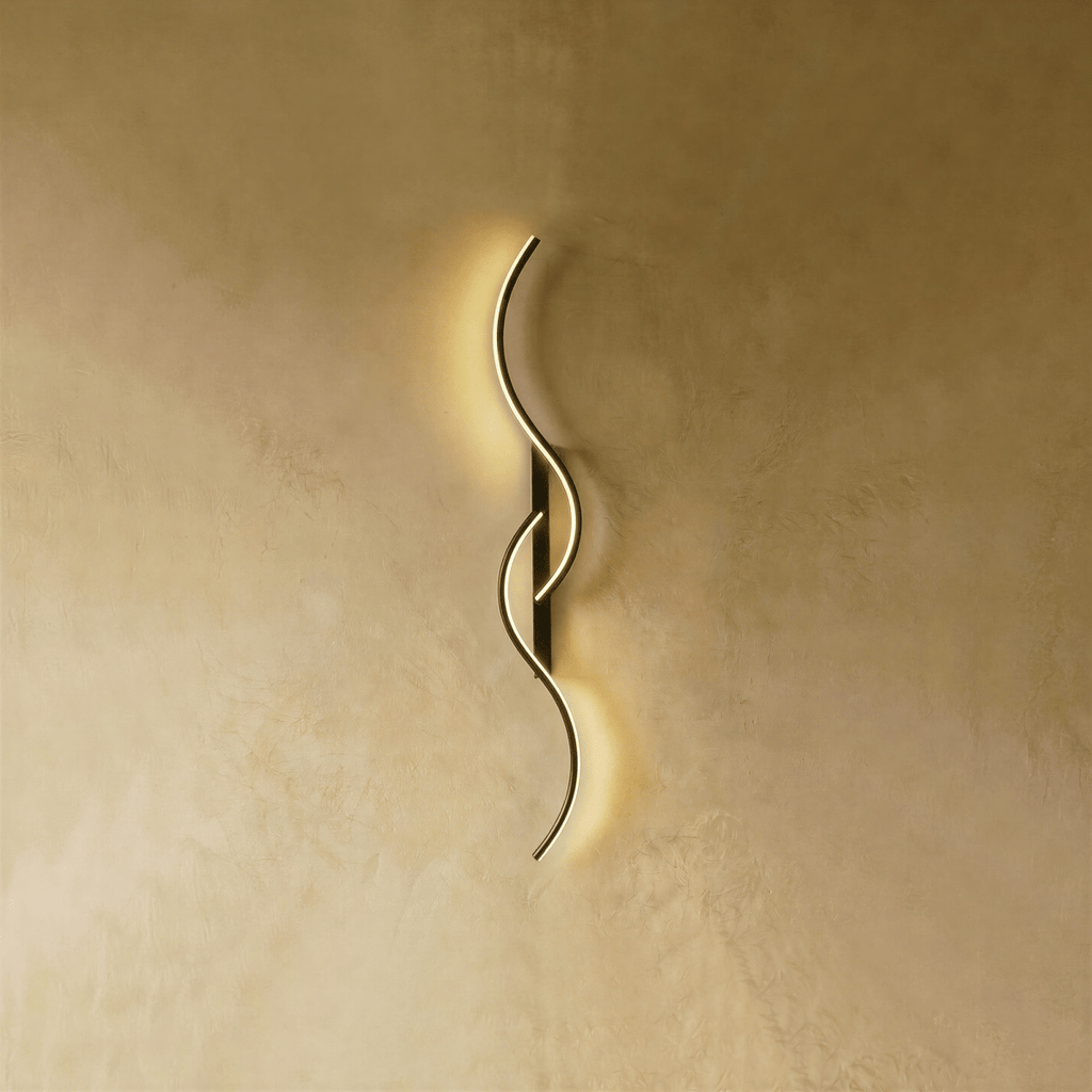 Clérachic S-shaped wall light with fine gold LED curves on a black backplate, shown lit with a soft, even glow on a warm wall.