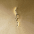 Clérachic S-shaped wall light with fine gold LED curves on a black backplate, shown lit with a soft, even glow on a warm wall.