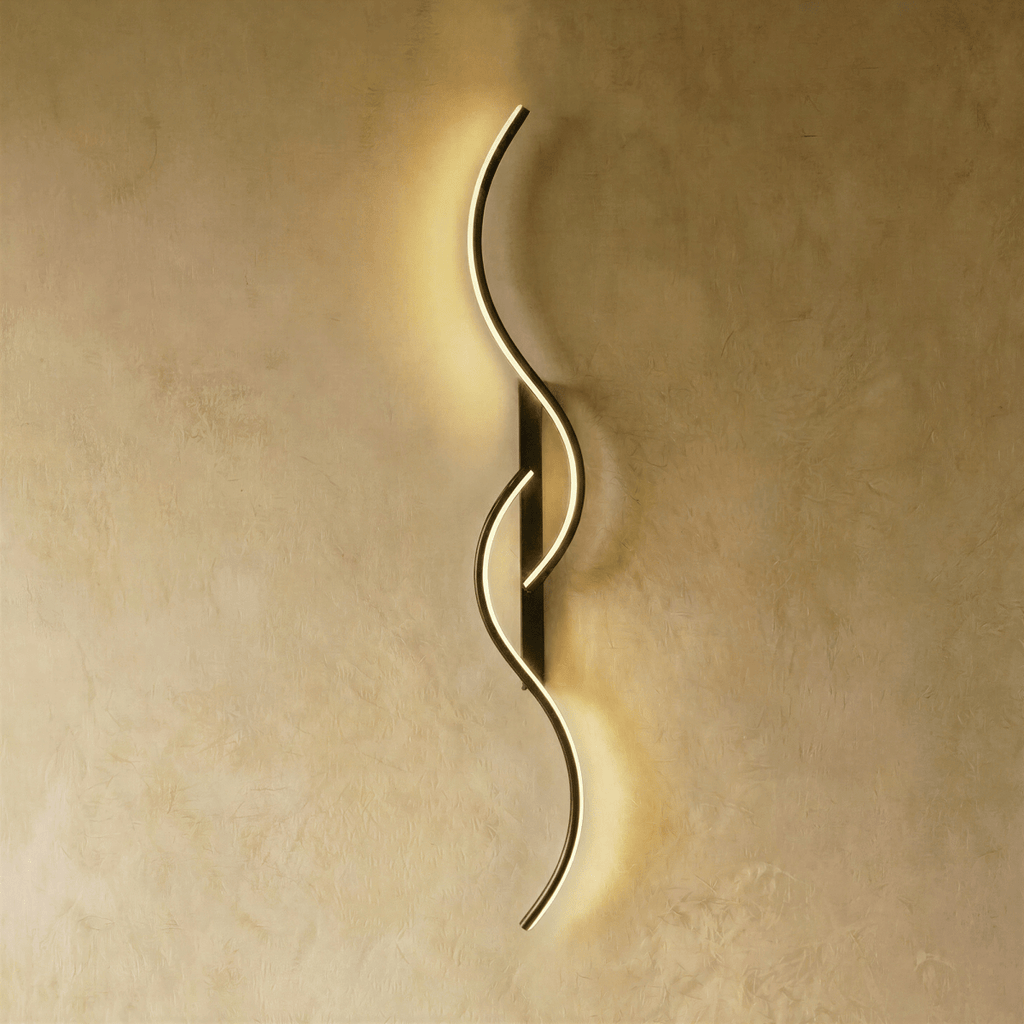 Clérachic tall black S-shaped wall light with two overlapping LED curves, casting a gentle warm glow down a plaster wall.