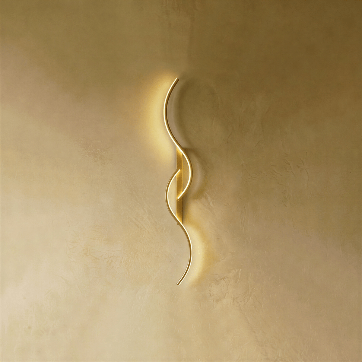 Clérachic gold S-shaped wall light with two interlocking LED curves and a matching gold backplate, glowing on warm plaster.