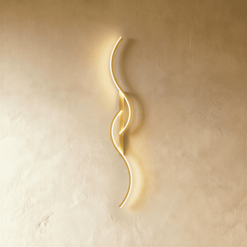 Clérachic gold S-shaped wall light, curves and slim backplate in one brushed gold finish, lit softly against pale plaster.