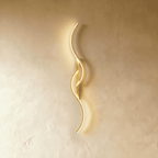 Clérachic gold S-shaped wall light with linked LED curves and a narrow gold bar, giving a soft warm glow on a plaster wall.