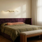 Clérachic gold S-shaped wall light mounted horizontally above a plum velvet headboard in a calm bedroom with sheer curtains.