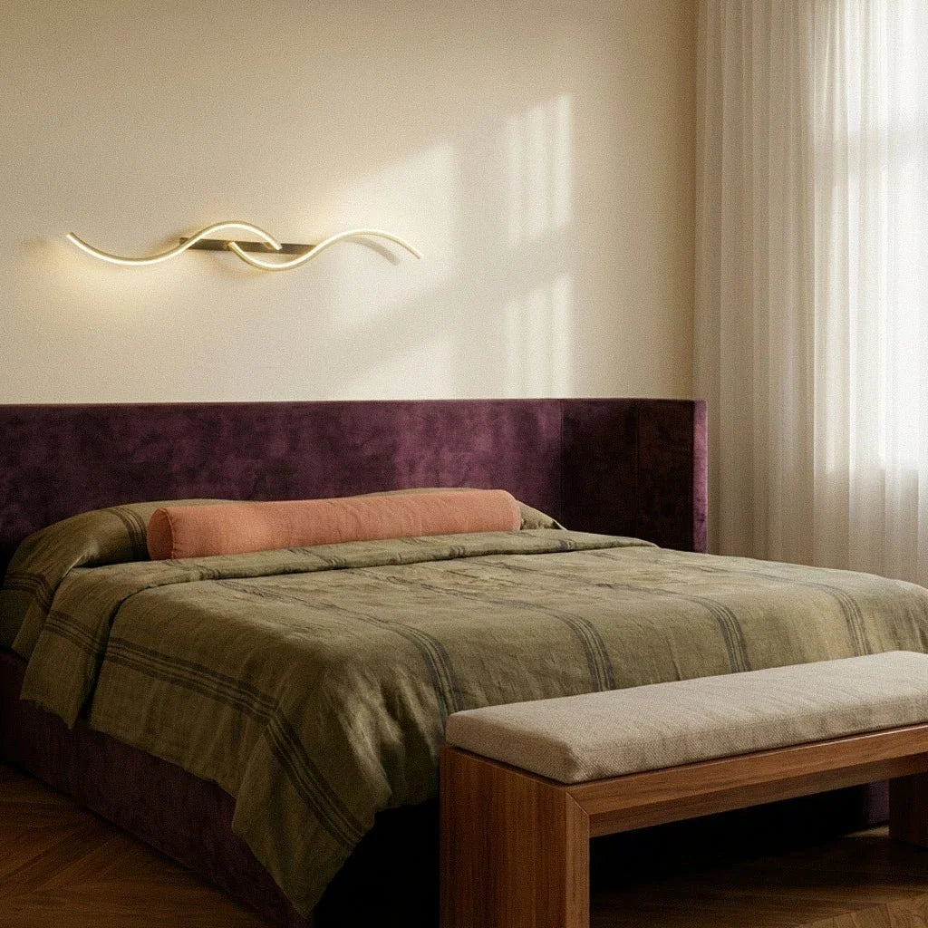 Clérachic gold S-shaped wall light mounted horizontally above a plum velvet headboard in a calm bedroom with sheer curtains.