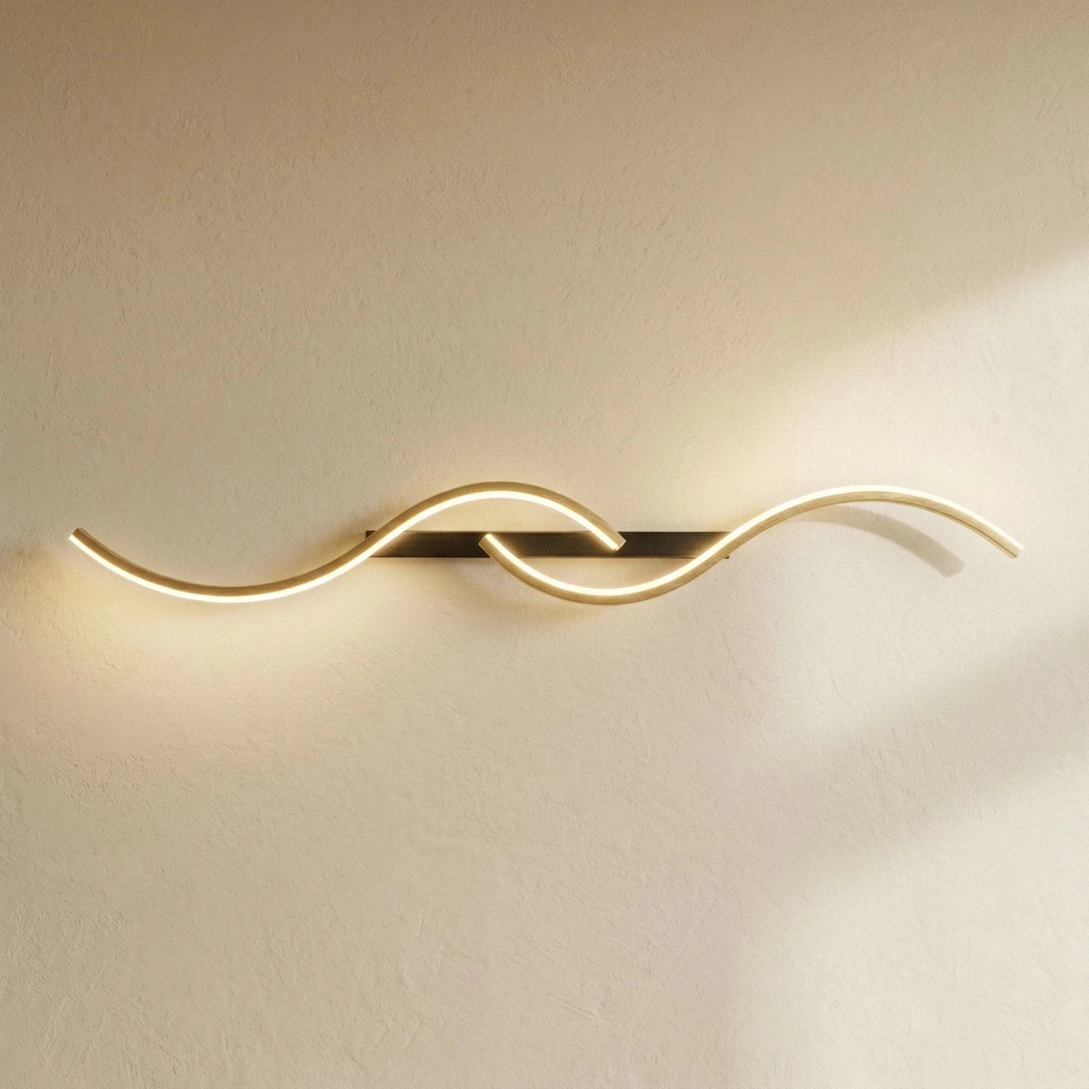 Clérachic S-shaped wall light turned horizontal, with gold LED curves on a black bar, glowing against a sunlit plaster wall.