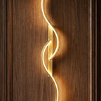 Close view of a Clérachic gold S-shaped wall light, showing the brushed gold backplate and glowing LED curve on walnut panelling.