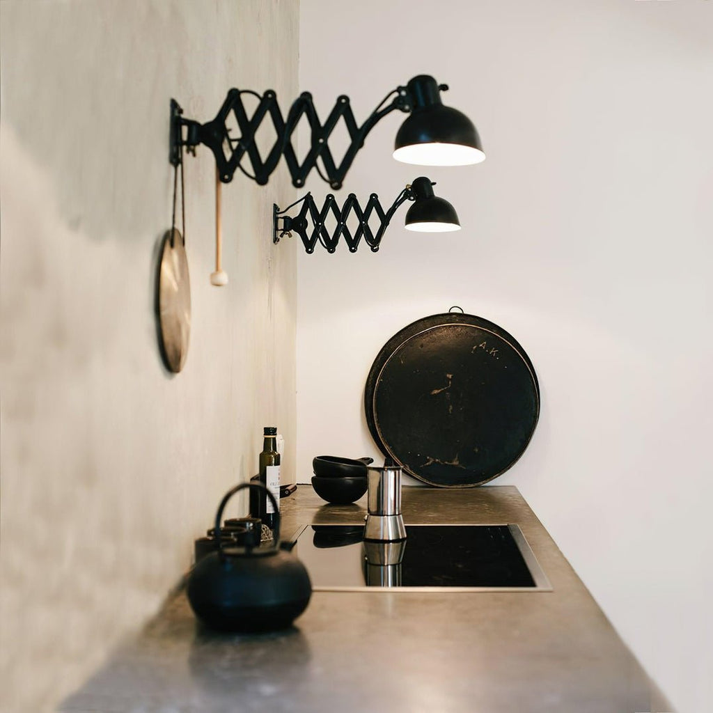 Two black Clérachic scissor-arm wall lights above a kitchen worktop with a hob, an iron kettle and a large black pan.