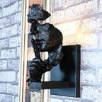 Side view of a black Clérachic wall light, the sculpted face and raised finger standing out from a black plate on a pale brick wall.