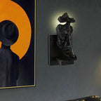 Black Clérachic face wall light on a dark grey wall, with light glowing from the eye gap and an orange framed painting to the left.