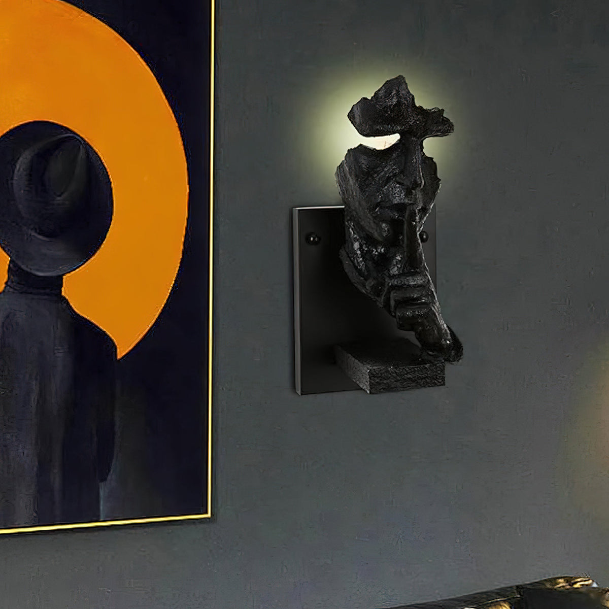 Black Clérachic face wall light on a dark grey wall, with light glowing from the eye gap and an orange framed painting to the left.