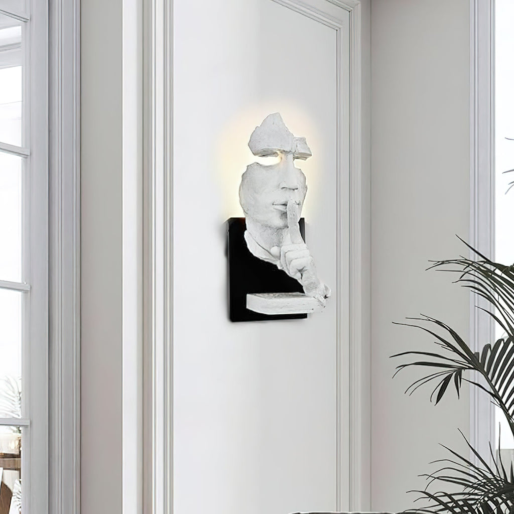 White Clérachic wall light with the sculpted face lit from behind, mounted on a white wall beside a tall window and a palm.