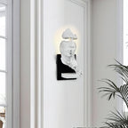 White Clérachic wall light with the sculpted face lit from behind, mounted on a white wall beside a tall window and a palm.