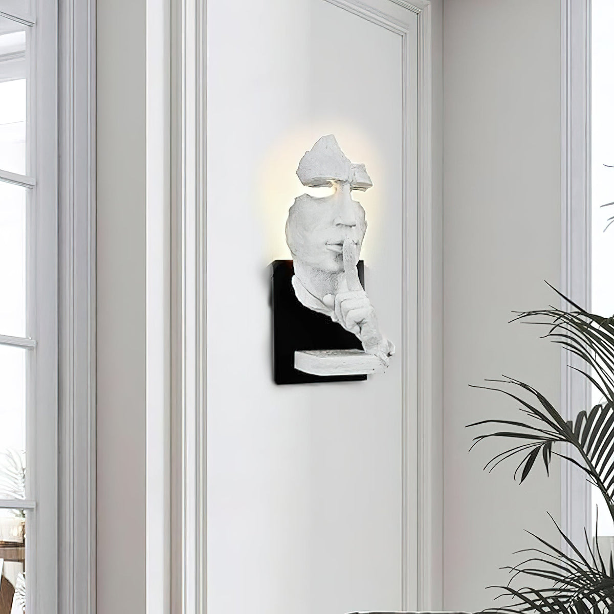 White Clérachic wall light with the sculpted face lit from behind, mounted on a white wall beside a tall window and a palm.