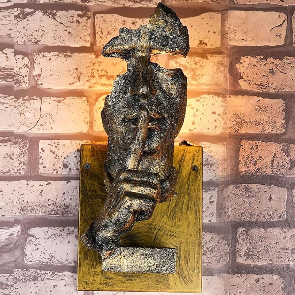 A bronze-finish Clérachic wall light on a rustic brick wall, warm light glowing through the face and a worn gold-toned backplate.