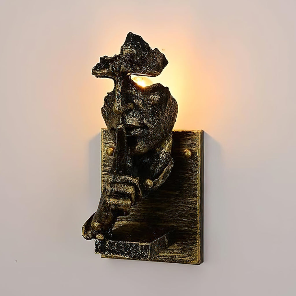 Bronze Clérachic face wall light on a soft peach wall, light shining from the eye gap above a finger raised to the lips.