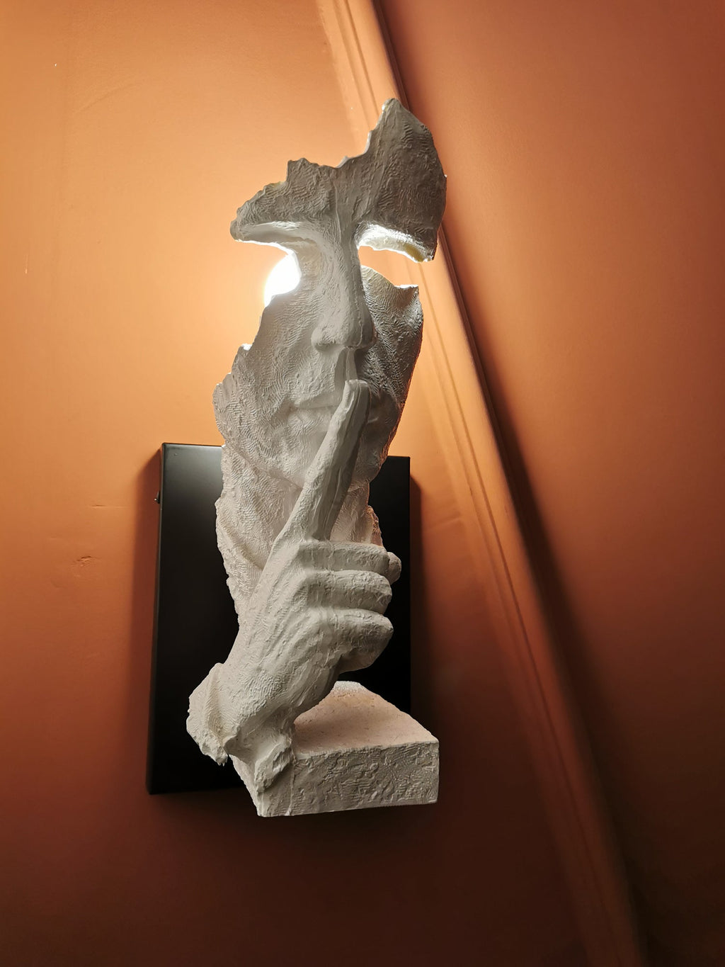 Another close view of the white Clérachic face wall light, glowing in the eye gap against an orange wall.