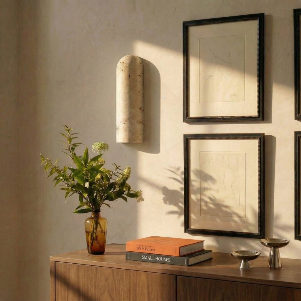 Clérachic travertine wall light mounted above a walnut sideboard with a vase of foliage, framed prints and books in soft sunlight.