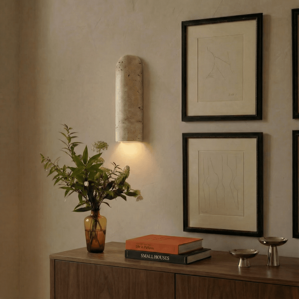 Clérachic travertine wall light glowing warmly at dusk above a walnut sideboard, beside framed line drawings and an amber glass vase.