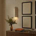 Clérachic travertine wall light glowing warmly at dusk above a walnut sideboard, beside framed line drawings and an amber glass vase.