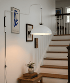 This white Clérachic wall light hangs over a staircase landing with framed prints and a small plant on a wooden stool.