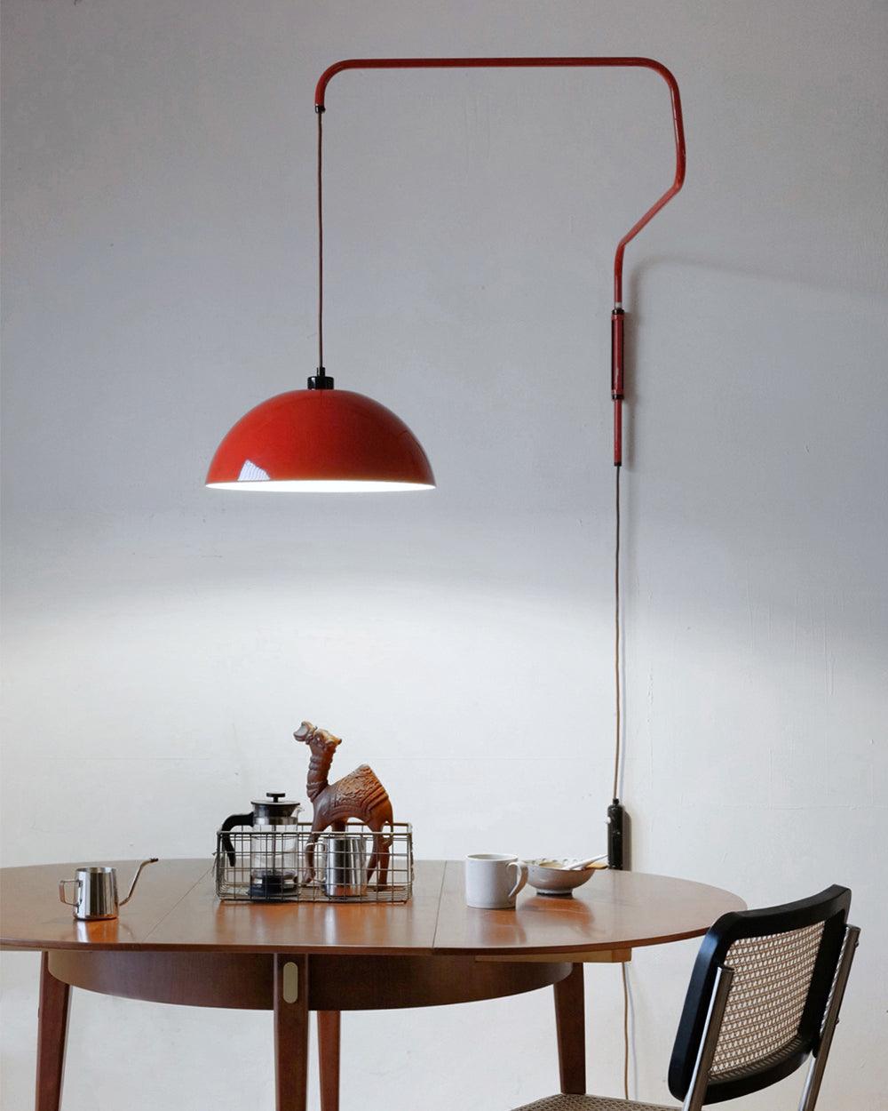 Red dome and swing arm of this Clérachic wall light above a round wooden table with a small wooden camel on top.