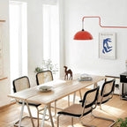 A red Clérachic wall light seen over a long pale wooden dining table with black cane-back chairs and a Matisse-style print.
