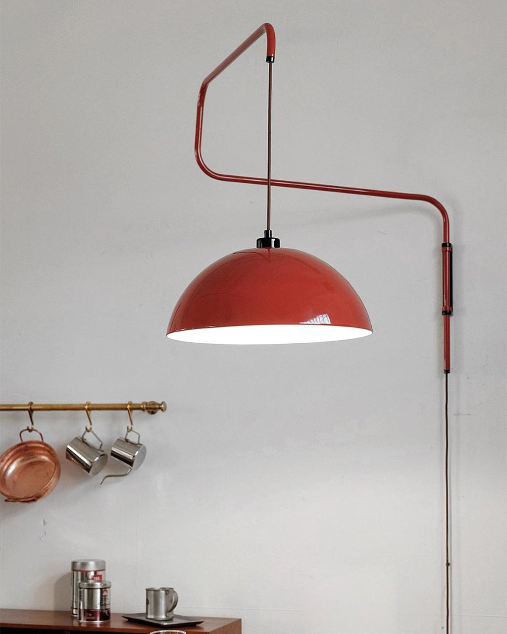 Glossy red shade of this Clérachic wall light on a grey wall, above a rail of copper pans and mugs.