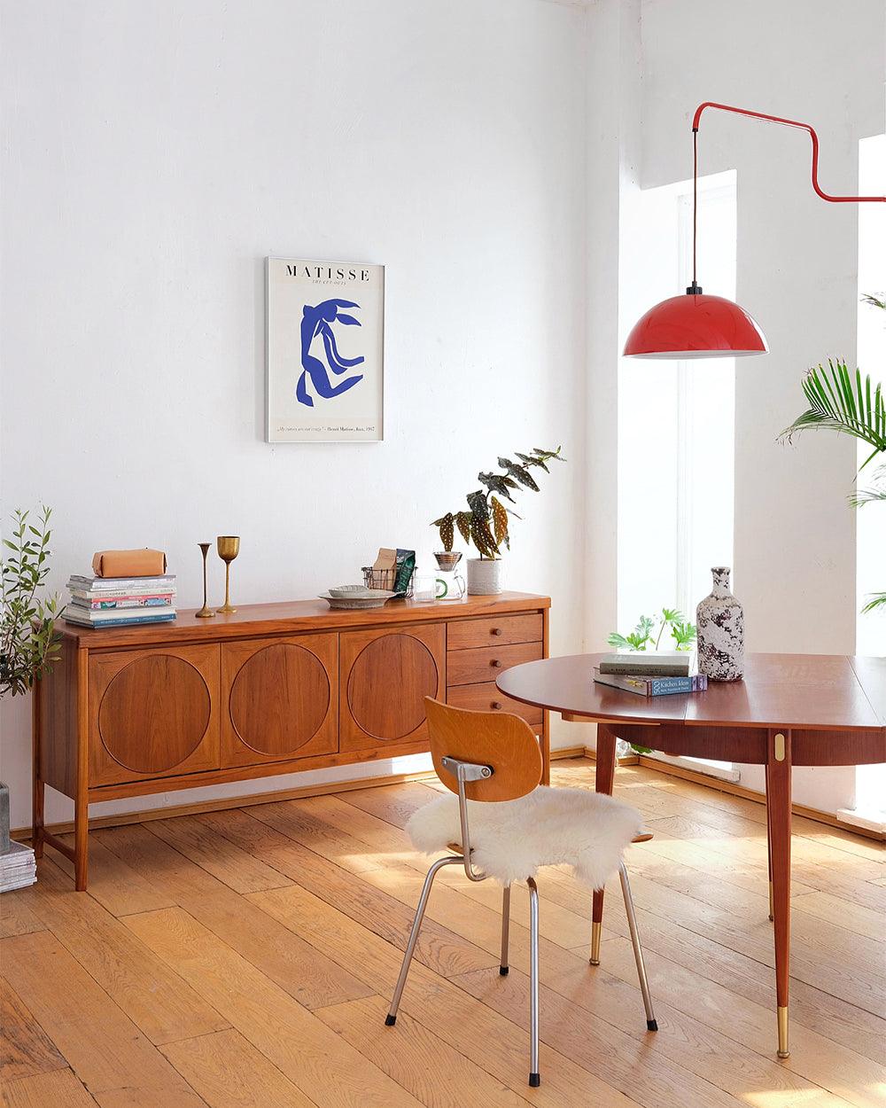 This Clérachic wall light in red hangs in a bright room with a wooden sideboard, a round table and a blue print.