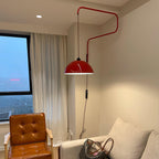 This Clérachic wall light in red beside a tall window and cream curtains, above a tan leather armchair and a grey sofa.