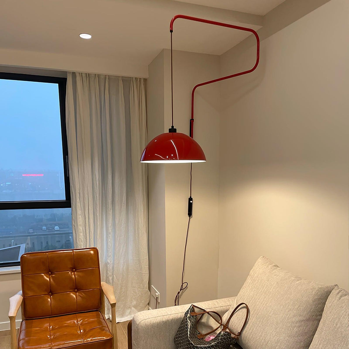This Clérachic wall light in red beside a tall window and cream curtains, above a tan leather armchair and a grey sofa.