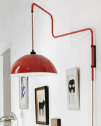 A red Clérachic wall light hangs over a pale wall, with a spiral stem and a cable hanging down from its black switch.