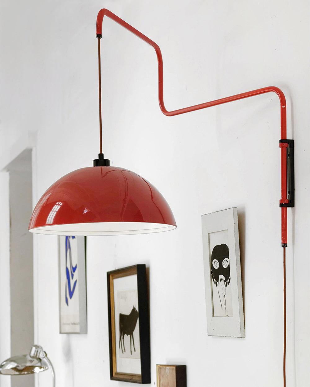 A red Clérachic wall light hangs over a pale wall, with a spiral stem and a cable hanging down from its black switch.