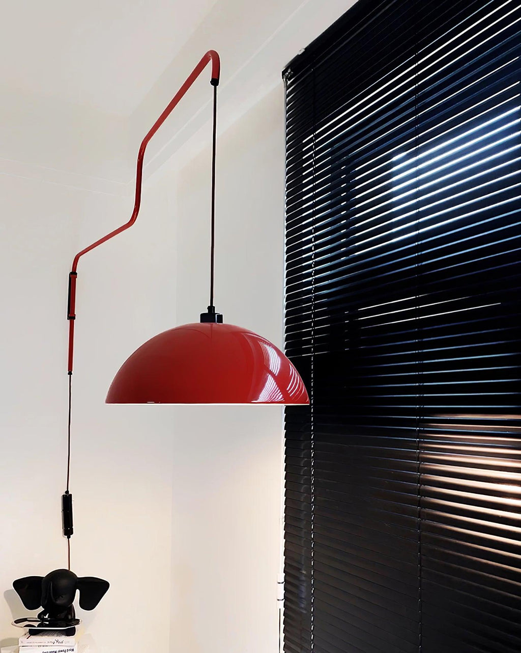 A red Clérachic wall light hangs near black window blinds, its cable running down past a small black ornament.