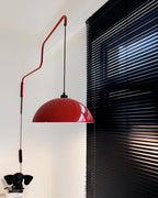 A red Clérachic wall light hangs near black window blinds, its cable running down past a small black ornament.