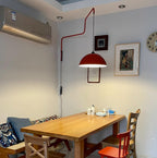 This Clérachic wall light in red hangs above a wooden dining table in a room with an air-conditioner and wall clock.