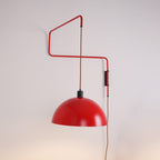 Red swing-arm wall light with a domed shade, this Clérachic wall light against a pale pink-grey wall, with a warm glow below.