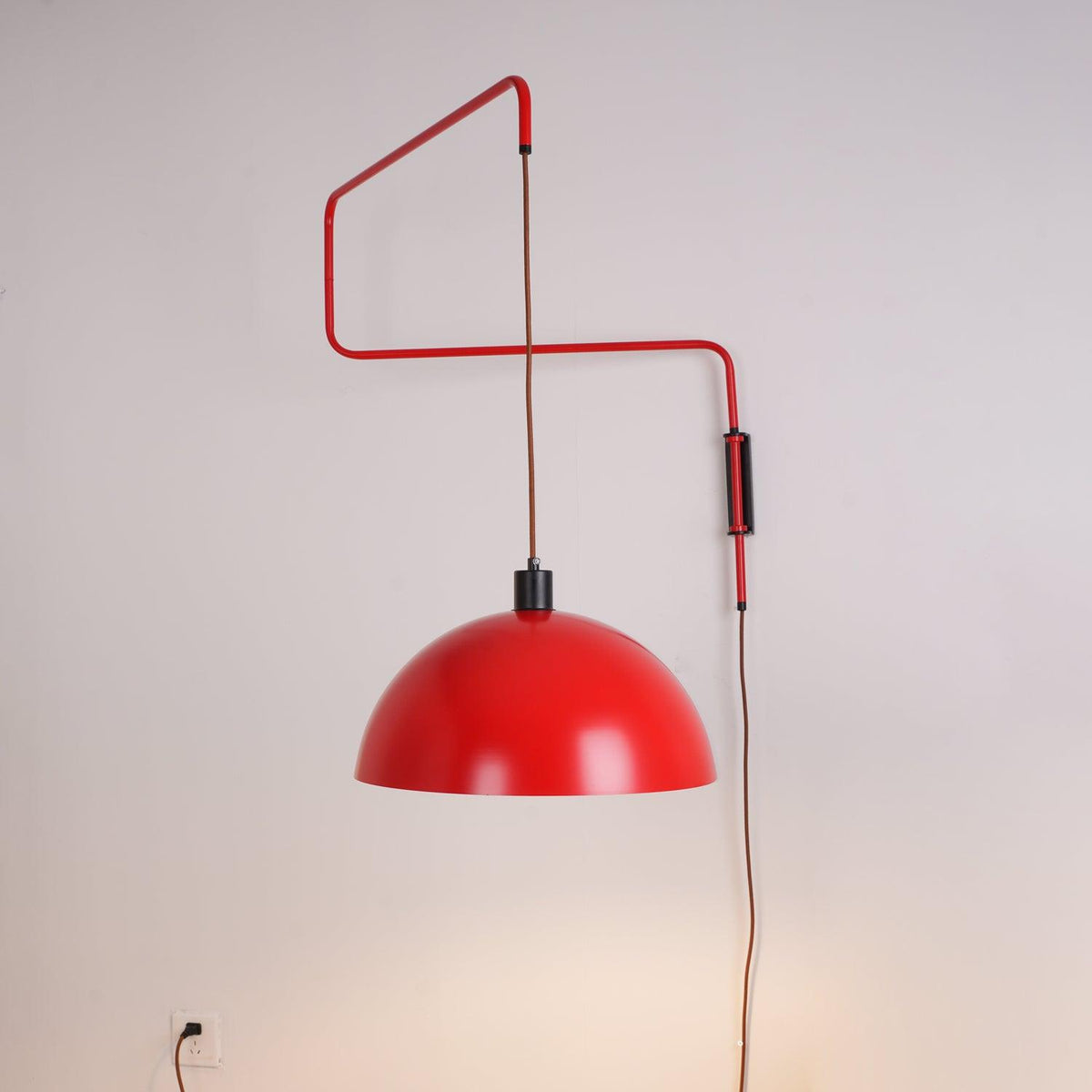 Zigzag arm and red shade of this Clérachic wall light on a soft grey wall, with a wall socket at lower left.