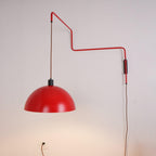 A red Clérachic wall light on a pale wall, the shade swung out to the left, with its cable running to a wall socket.