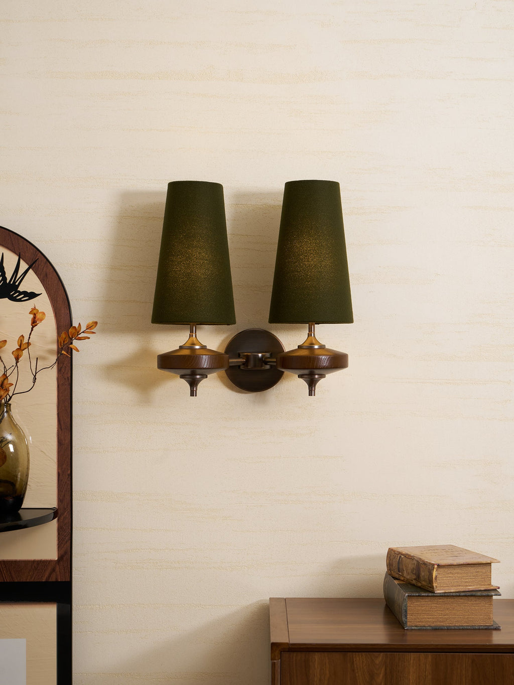 Twin dark-green shades glow softly on this Clérachic wall sconce, mounted on a cream wall beside an arched mirror and a walnut cabinet.