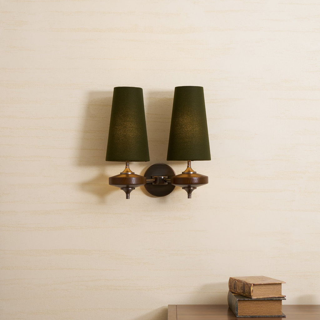 Two green tapered shades and walnut wood bases on a Clérachic wall light, hung on a pale textured wall above a stack of books.