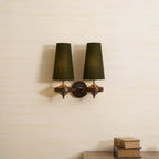 Two green tapered shades and walnut wood bases on a Clérachic wall light, hung on a pale textured wall above a stack of books.