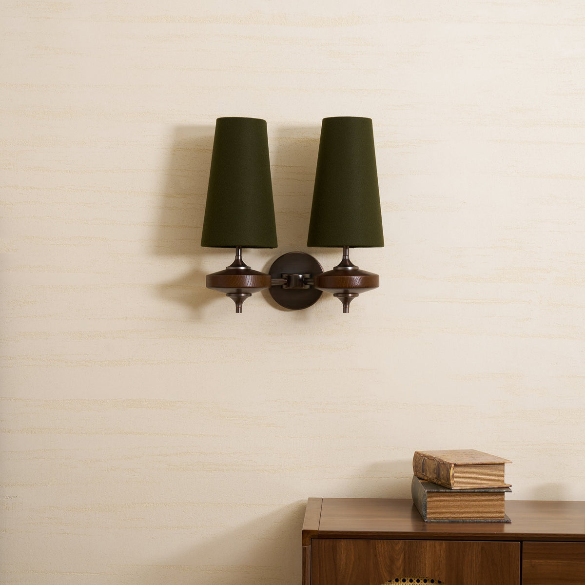 Close, straight-on view of a Clérachic twin-shade wall light with green fabric shades, bronze stems and walnut discs on a cream wall.