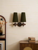 A Clérachic two-light sconce in green hangs beside an arched mirror, with a rattan-fronted cabinet and old books below.