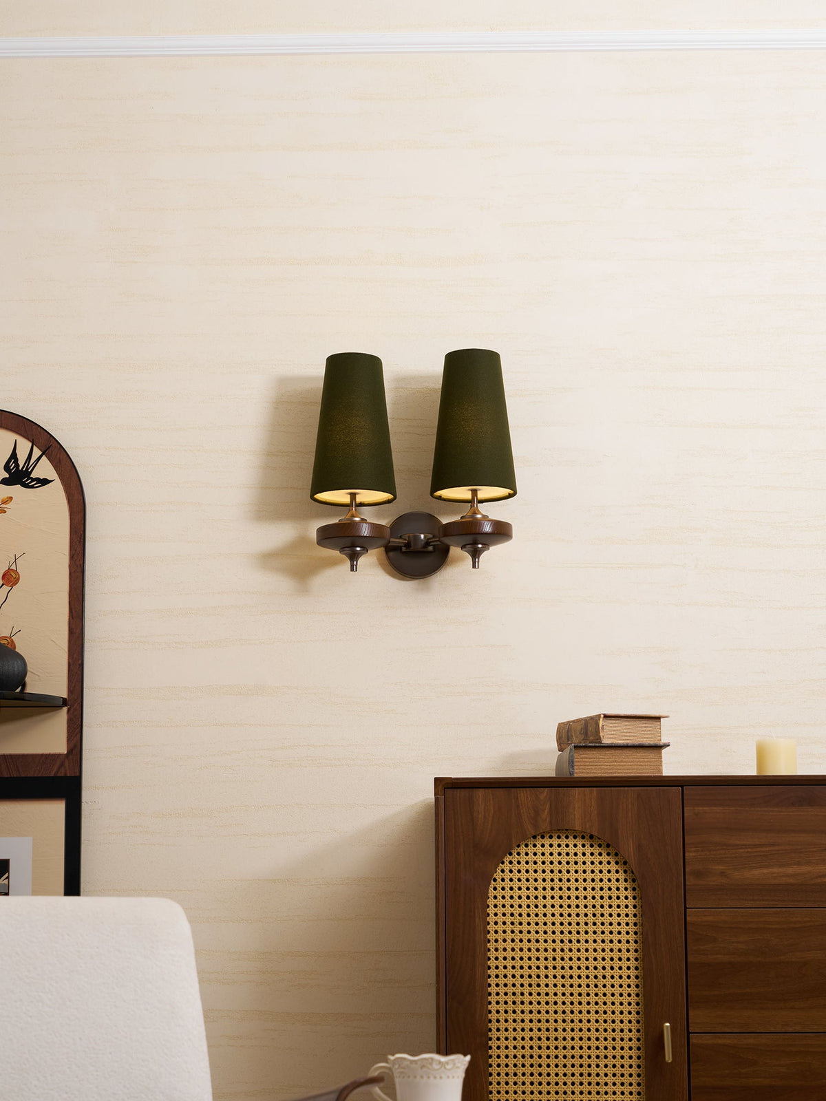 A Clérachic twin sconce with green shades lit warm from within, set above a rattan-panelled cabinet and a pale armchair.