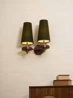 Lit green shades and brass-toned arms on a Clérachic wall light, mounted on a pale wall over a walnut cabinet and a pile of books.